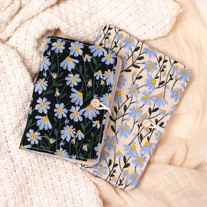 Peut inclure: Deux couvertures de livre brodées. L'une est noire avec des fleurs bleues et jaunes, l'autre est crème avec des fleurs bleues et jaunes. Les deux ont une fermeture à bouton en bois et une ficelle blanche. Les couvertures sont posées sur un tissu crème.