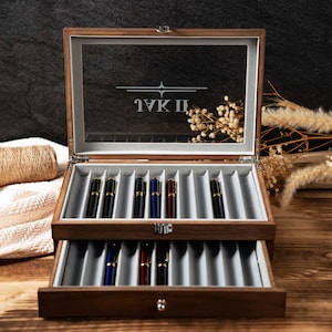 Op de afbeelding: Een houten pennen display case met een glazen deksel, met verschillende pennen in verschillende kleuren. De case heeft twee niveaus met individuele sleuven voor elke pen. De tekst "IVK II" staat op de bovenkant.