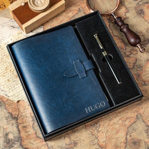 Peut inclure: Un ensemble cadeau comprenant un journal en cuir bleu avec une fermeture, personnalisé avec le nom "HUGO", et un stylo noir avec des détails dorés, présenté dans un coffret noir. L'ensemble est posé sur une carte vintage.
