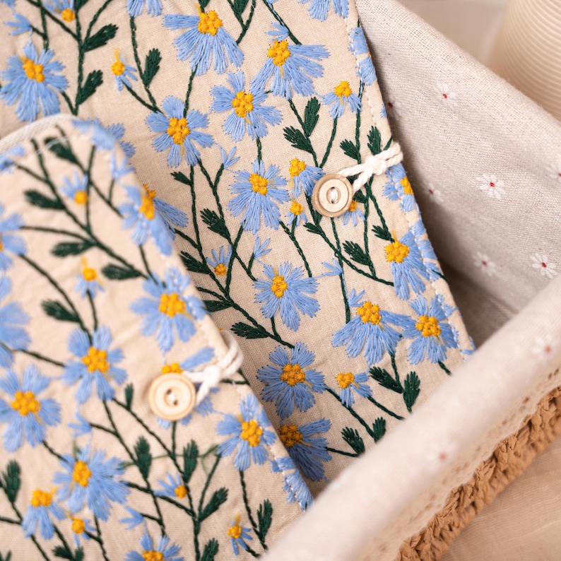 May include: Close-up of two beige fabric items with blue daisy embroidery and wooden button closures. The daisies have yellow centers and green stems. The items are inside a woven basket.