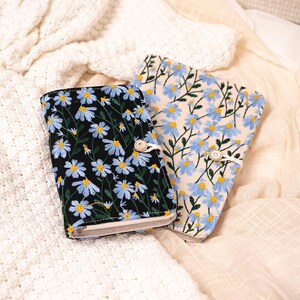 May include: Two embroidered fabric journals. One journal has a black background with blue and yellow daisy flowers. The other journal has a cream background with blue and yellow daisy flowers. Both journals have a button closure.