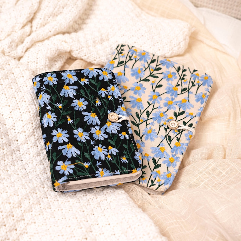 May include: Two embroidered fabric journals. One journal has a black background with blue and yellow daisy flowers. The other journal has a cream background with blue and yellow daisy flowers. Both journals have a button closure.