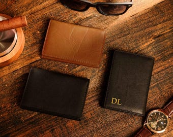 May include: Three leather wallets on a wooden surface. One wallet is brown, one is black, and one is black with the initials "DL" embossed in gold.