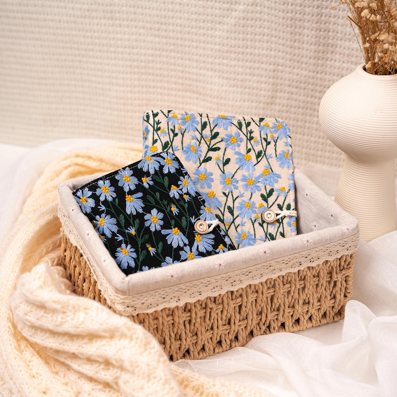May include: Two floral embroidered notebooks in a woven basket. One notebook has a black background with blue flowers, the other has a cream background with blue flowers. The basket is lined with cream fabric and lace trim.
