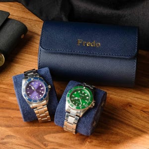 Peut inclure: Deux montres-bracelets argentées avec des cadrans bleu et vert sont présentées sur des coussins en velours bleu. Les montres sont dans un porte-montres en cuir bleu avec le mot "Fredo" embossé en or.
