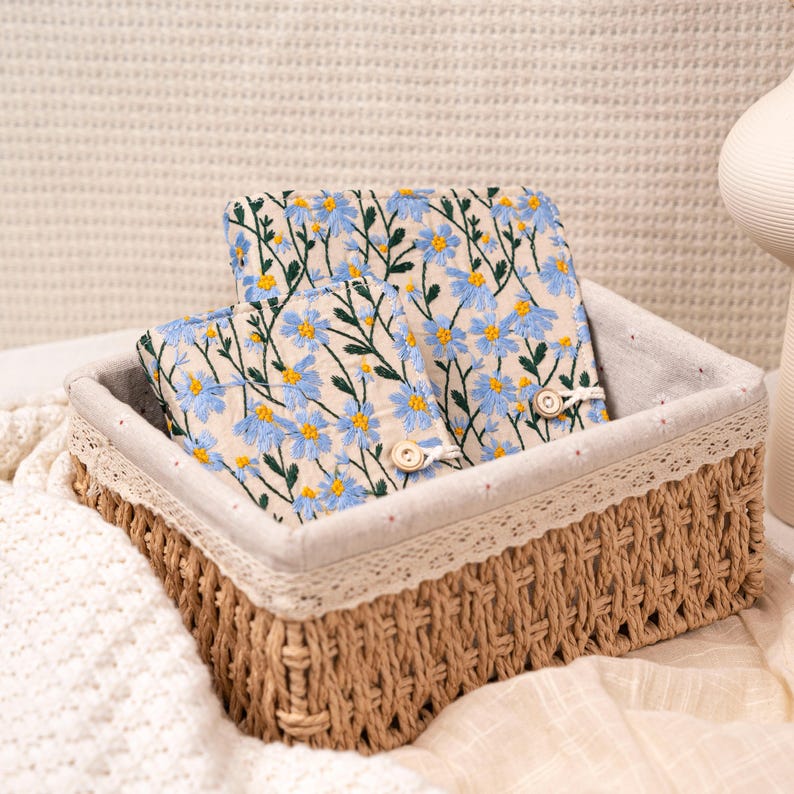 May include: A woven basket with a beige interior and a decorative lace trim. Inside the basket are two embroidered fabric items with a pattern of blue flowers and green stems. The basket is sitting on a white surface.
