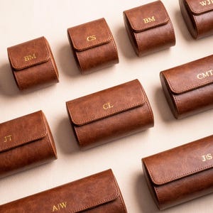 May include: Ten brown leather watch roll cases with gold lettering. Each case has a different two-letter monogram.
