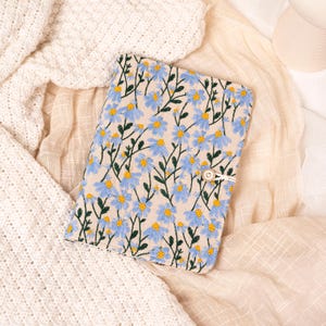 May include: A beige journal cover with embroidered blue flowers and green stems. The cover is secured with a button and a loop closure. The journal is lying on a cream-colored fabric.