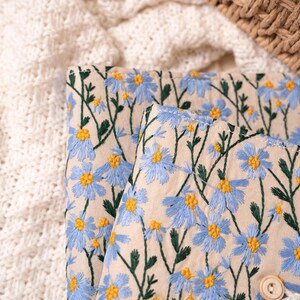 May include: Close-up of a beige fabric with embroidered blue daisies and yellow centers. Green stems and leaves are also embroidered. A white crocheted blanket and a woven basket are in the background.