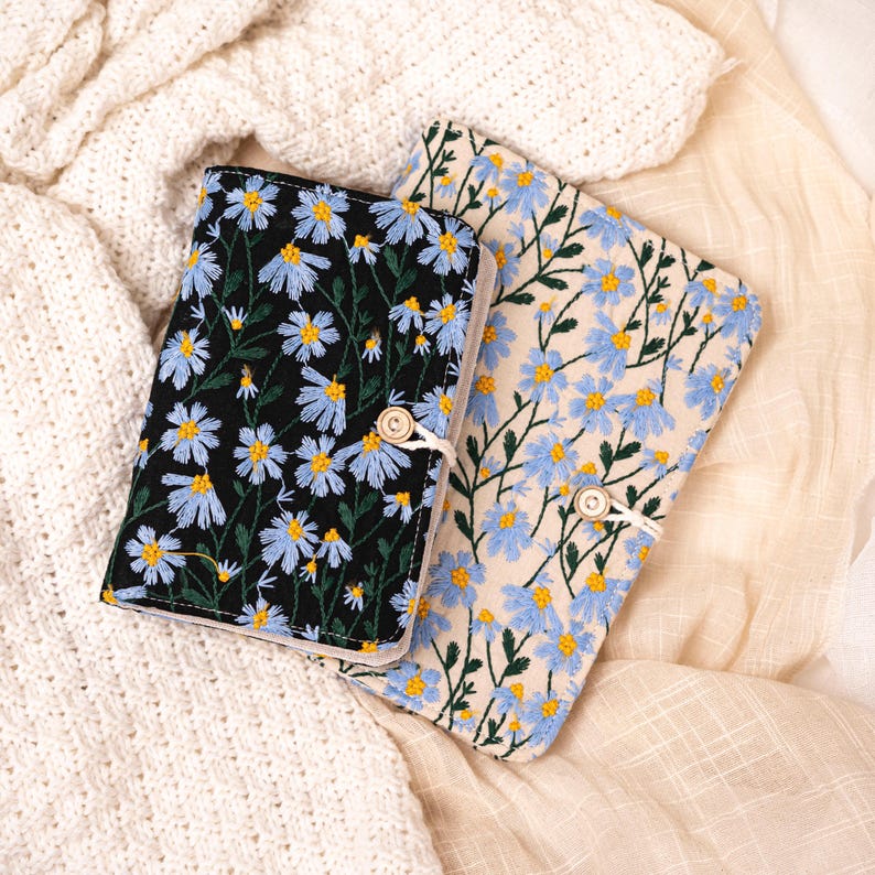 May include: Two embroidered book covers. One is black with blue and yellow flowers, the other is cream with blue and yellow flowers. Both have a wooden button closure and white string. The covers are lying on a cream-colored fabric.