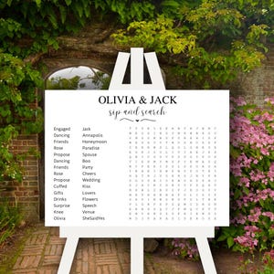 May include: A white sign with black text that says "Olivia & Jack sip and search". The sign has a word search puzzle with words related to weddings, such as "engaged", "wedding", "honeymoon", "love", and "venue".