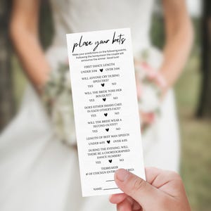 May include: A white wedding game card with the text "place your bets" in elegant script. The card lists questions about the wedding day, such as the first dance length and if the bride will toss her bouquet. The card is held by a person.