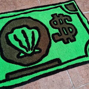 以下が含まれることがあります： 黒い輪郭のあるドル札の形をした緑色の敷物。敷物の中央には茶色の貝殻と黒いドルマークがあります。