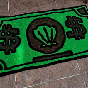 以下が含まれることがあります： 黒色の輪郭で囲まれた、ドル紙幣の形をした緑色の敷物。敷物の中央には、黒と白の大きな貝殻のデザインがあり、両側にドル記号が2つある。