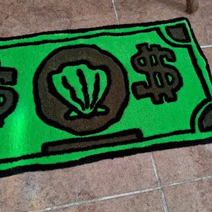 以下が含まれることがあります： 黒の輪郭と中央の茶色の貝殻を持つ、ドル札の形をした緑色の敷物。敷物は右側に黒いドル記号があります。