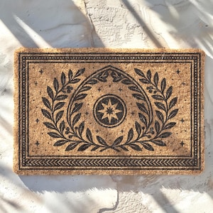 May include: Rectangular doormat made of natural coir fiber. The mat features a black decorative border and a central design of a laurel wreath surrounding a starburst. The design is symmetrical and detailed, suitable for a home entrance.