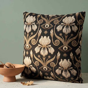 May include: A square throw pillow featuring a black fabric base with an intricate embroidered pattern in gold and beige thread. The design consists of repeating iris flowers, stylized eyes, and small star shapes arranged in a symmetrical layout.