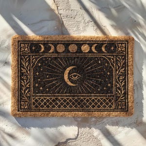 Pode incluir: Capacho retangular de fibra de coco com fundo castanho escuro e design celestial. O design apresenta uma lua crescente com um olho, raios de sol, estrelas e fases da lua. A borda tem padrões florais e geométricos.