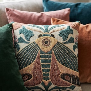 May include: A square throw cushion featuring a detailed embroidered moth design in shades of teal, mustard yellow, and rust brown on a cream-coloured background. The moth is centred with symmetrical wings and intricate patterns, surrounded by floral motifs.