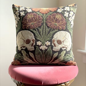 Op de afbeelding: Vierkantig sierkussen met een donkere achtergrond met twee schedels en bloemmotieven in bruin-, groen- en wittinten. Het kussen ligt op een roze fluwelen stoel.