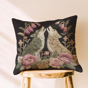 A square woven tapestry pillow with two howling wolves on a black background. The wolves are surrounded by pink and green botanical flowers, creating a vintage, folkloric scene. This decorative cushion is perfect for cottagecore, dark academia, and gothic home decor.