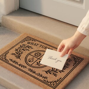 Natural coir doormat featuring "What Turns, Returns" text with sacred elemental symbolism including sun, crescent moon, flames, and botanical sprigs arranged in protective circular mandala design. Four classical elements represented in quadrants surrounded by intricate corner flourishes and leafy botanical borders. Perfect witchy welcome mat for pagan home decor, spiritual protection, and mystical entryway styling.