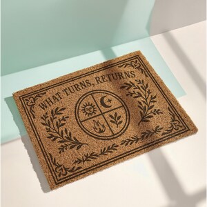 Natural coir doormat featuring "What Turns, Returns" text with sacred elemental symbolism including sun, crescent moon, flames, and botanical sprigs arranged in protective circular mandala design. Four classical elements represented in quadrants surrounded by intricate corner flourishes and leafy botanical borders. Perfect witchy welcome mat for pagan home decor, spiritual protection, and mystical entryway styling.