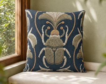 May include: A square throw cushion featuring a dark blue fabric base with an embroidered olive green beetle at the centre. The design includes symmetrical, stylised floral and leaf patterns in muted beige and green tones surrounding the insect.