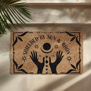 May include: A brown coir doormat with the text "Shielded by Sun & Moon" in black lettering. The design features two hands reaching up towards a crescent moon and sun with stars.