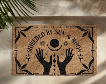 May include: A brown coir doormat with the text "Shielded by Sun & Moon" in black lettering. The design features two hands reaching up towards a crescent moon and sun with stars.