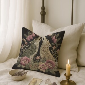 A square woven tapestry pillow with two howling wolves on a black background. The wolves are surrounded by pink and green botanical flowers, creating a vintage, folkloric scene. This decorative cushion is perfect for cottagecore, dark academia, and gothic home decor.