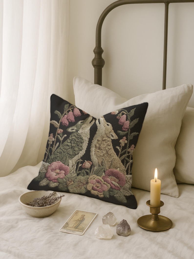 A square woven tapestry pillow with two howling wolves on a black background. The wolves are surrounded by pink and green botanical flowers, creating a vintage, folkloric scene. This decorative cushion is perfect for cottagecore, dark academia, and gothic home decor.