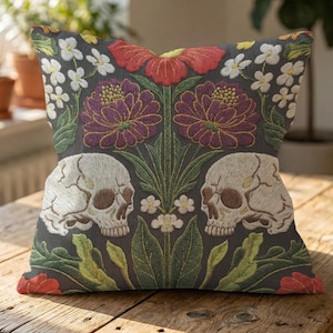 Grimoire and Goblet gothic pillow cover featuring mirrored ivory human skulls surrounded by detailed floral and botanical elements including deep red flowers, purple blooms with gold outlining, small white blossoms, and layered green foliage, arranged in a symmetrical composition on a charcoal background, designed with a high-detail faux embroidery print that mimics stitched texture while remaining smooth, creating a dark academia and gothic botanical aesthetic decorative cushion cover