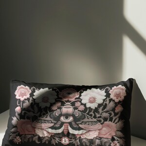 Gothic moth eye botanical lumbar pillow with mystical embroidered wings on dark fabric. Cottagecore throw pillow featuring protective moth symbol surrounded by pink roses, white wildflowers & sacred botanicals. Witchy home decor with medieval tapestry aesthetic. Perfect for dark academia spaces, bohemian decor & occult art lovers. 20x12 lumbar size.