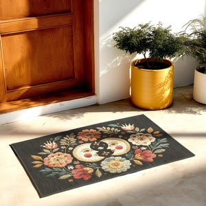 Witchy outdoor doormat featuring a central all-seeing eye, crescent moons, and floral details in an embroidery-style design. This weatherproof welcome mat measures 26x18 inches and is ideal for porches or patio entries. Symbolic celestial motifs offer intuitive protection and spiritual elegance for magical home decor.