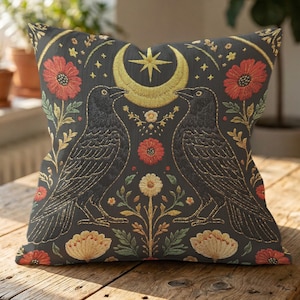 May include: Decorative throw pillow with a dark gray background featuring embroidered black ravens facing each other. The design includes a gold crescent moon, star, and floral accents in red, yellow, and green.