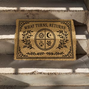 Natural coir doormat featuring "What Turns, Returns" text with sacred elemental symbolism including sun, crescent moon, flames, and botanical sprigs arranged in protective circular mandala design. Four classical elements represented in quadrants surrounded by intricate corner flourishes and leafy botanical borders. Perfect witchy welcome mat for pagan home decor, spiritual protection, and mystical entryway styling.