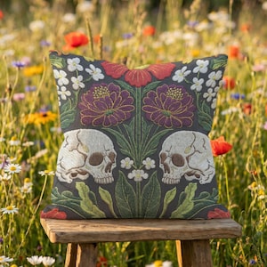 Grimoire and Goblet gothic pillow cover featuring mirrored ivory human skulls surrounded by detailed floral and botanical elements including deep red flowers, purple blooms with gold outlining, small white blossoms, and layered green foliage, arranged in a symmetrical composition on a charcoal background, designed with a high-detail faux embroidery print that mimics stitched texture while remaining smooth, creating a dark academia and gothic botanical aesthetic decorative cushion cover