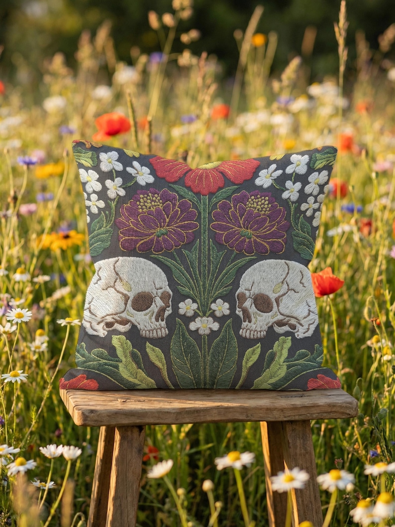 Grimoire and Goblet gothic pillow cover featuring mirrored ivory human skulls surrounded by detailed floral and botanical elements including deep red flowers, purple blooms with gold outlining, small white blossoms, and layered green foliage, arranged in a symmetrical composition on a charcoal background, designed with a high-detail faux embroidery print that mimics stitched texture while remaining smooth, creating a dark academia and gothic botanical aesthetic decorative cushion cover