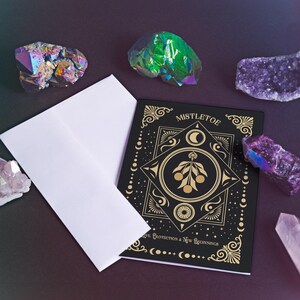 Luxurious black Yule card featuring golden mistletoe design, moon phases, and stars. "Mistletoe" and "For Protection & New Beginnings" in ornate gold text.