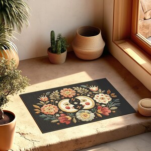 Witchy outdoor doormat featuring a central all-seeing eye, crescent moons, and floral details in an embroidery-style design. This weatherproof welcome mat measures 26x18 inches and is ideal for porches or patio entries. Symbolic celestial motifs offer intuitive protection and spiritual elegance for magical home decor.