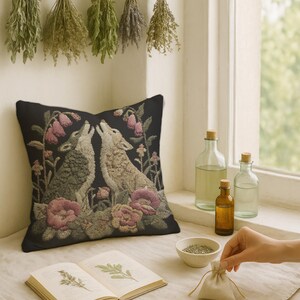 A square woven tapestry pillow with two howling wolves on a black background. The wolves are surrounded by pink and green botanical flowers, creating a vintage, folkloric scene. This decorative cushion is perfect for cottagecore, dark academia, and gothic home decor.