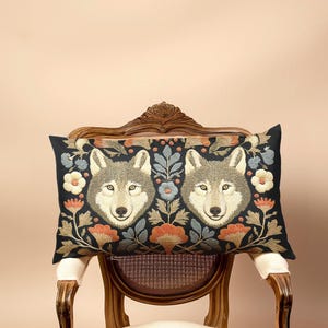 May include: A rectangular decorative pillow featuring two wolf faces surrounded by floral embroidery. The pillow has a black background with orange, blue, and white flowers. The pillow is placed on a vintage wooden chair.