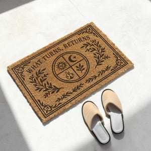 Natural coir doormat featuring "What Turns, Returns" text with sacred elemental symbolism including sun, crescent moon, flames, and botanical sprigs arranged in protective circular mandala design. Four classical elements represented in quadrants surrounded by intricate corner flourishes and leafy botanical borders. Perfect witchy welcome mat for pagan home decor, spiritual protection, and mystical entryway styling.