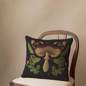 A dark woven pillow with a detailed embroidered mushroom and botanical design in deep red, green, and gold tones. Made with a cotton-poly blend.