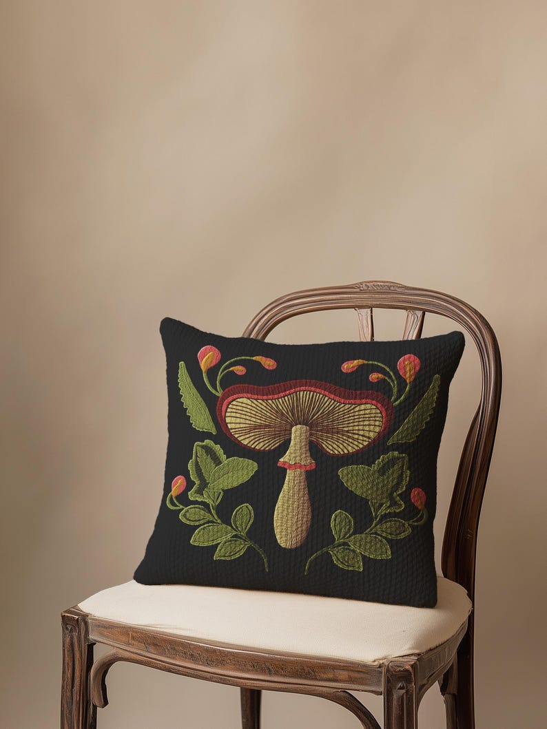 A dark woven pillow with a detailed embroidered mushroom and botanical design in deep red, green, and gold tones. Made with a cotton-poly blend.