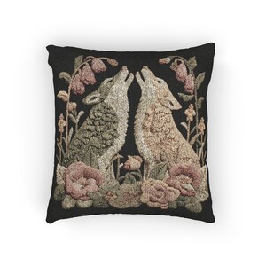 A square woven tapestry pillow with two howling wolves on a black background. The wolves are surrounded by pink and green botanical flowers, creating a vintage, folkloric scene. This decorative cushion is perfect for cottagecore, dark academia, and gothic home decor.