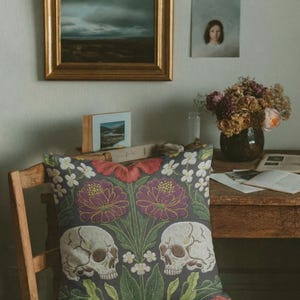 Grimoire and Goblet gothic pillow cover featuring mirrored ivory human skulls surrounded by detailed floral and botanical elements including deep red flowers, purple blooms with gold outlining, small white blossoms, and layered green foliage, arranged in a symmetrical composition on a charcoal background, designed with a high-detail faux embroidery print that mimics stitched texture while remaining smooth, creating a dark academia and gothic botanical aesthetic decorative cushion cover