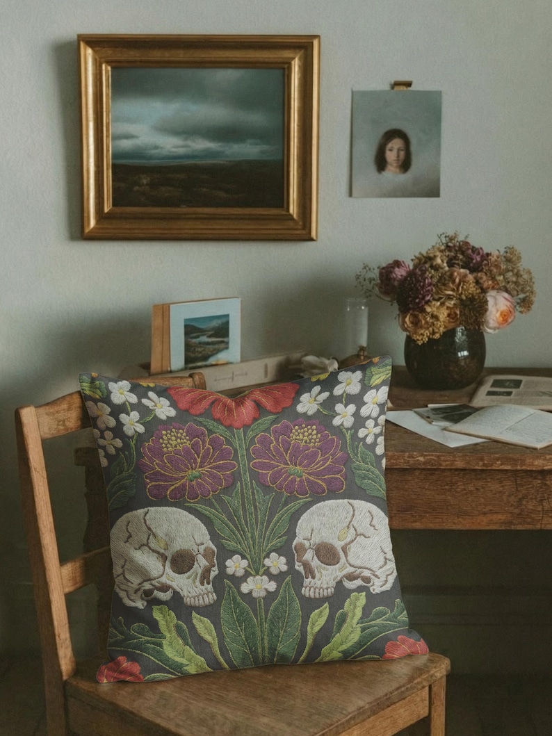 Grimoire and Goblet gothic pillow cover featuring mirrored ivory human skulls surrounded by detailed floral and botanical elements including deep red flowers, purple blooms with gold outlining, small white blossoms, and layered green foliage, arranged in a symmetrical composition on a charcoal background, designed with a high-detail faux embroidery print that mimics stitched texture while remaining smooth, creating a dark academia and gothic botanical aesthetic decorative cushion cover