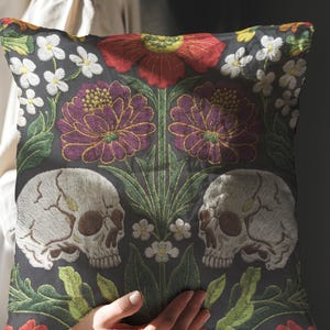 Grimoire and Goblet gothic pillow cover featuring mirrored ivory human skulls surrounded by detailed floral and botanical elements including deep red flowers, purple blooms with gold outlining, small white blossoms, and layered green foliage, arranged in a symmetrical composition on a charcoal background, designed with a high-detail faux embroidery print that mimics stitched texture while remaining smooth, creating a dark academia and gothic botanical aesthetic decorative cushion cover